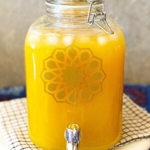 A clear glass jar filled with yellow-orange juice and ice sits on a checkered cloth. It features a decorative geometric pattern and a shiny silver spigot.