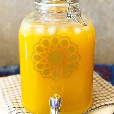 A clear glass jar filled with yellow-orange juice and ice sits on a checkered cloth. It features a decorative geometric pattern and a shiny silver spigot.