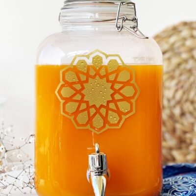 A glass drink dispenser filled with vibrant orange juice, featuring a silver spigot and decorative golden design, set on a colorful cloth.