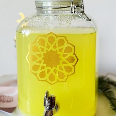 A glass jar filled with bright yellow lemonade, featuring an ornate gold geometric pattern. It has a metal spigot for pouring, conveying a refreshing and elegant atmosphere.