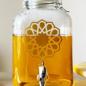 A glass jar with a silver spigot is filled with golden liquid. It features an intricate geometric pattern. The setting is bright and clean.