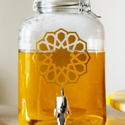 A glass jar with a silver spigot is filled with golden liquid. It features an intricate geometric pattern. The setting is bright and clean.