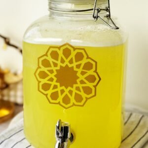 A clear glass dispenser filled with yellow lemonade, adorned with a gold geometric pattern. It sits on a striped cloth, conveying a fresh, cheerful vibe.