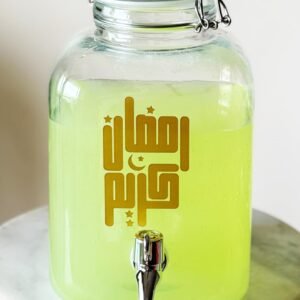 A clear glass dispenser filled with light green liquid on a marble stand. Arabic script and crescent moon on front; has a metal spout. Ramadan theme.