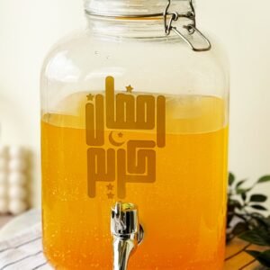 Glass jar with orange liquid, featuring Arabic text and stars. It's set on a striped cloth with a leafy plant nearby, evoking a festive vibe.