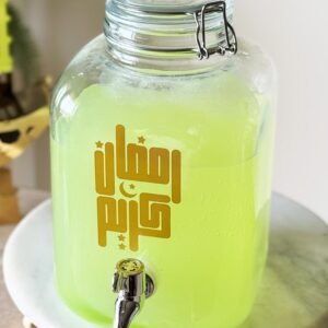 Glass jar with bright green liquid and Arabic text, featuring a metal spout, sits on a marble stand. The scene feels refreshing and inviting.