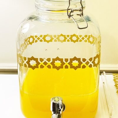 Large glass beverage dispenser filled with bright yellow liquid, adorned with a geometric gold pattern. It features a metal spigot, set on a white surface.