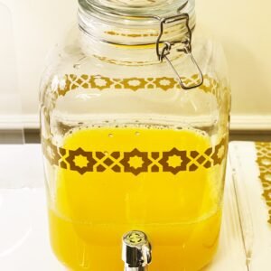 Glass beverage dispenser with yellow juice, sealed with a metal latch. It features a decorative geometric pattern and a chrome spigot, on a white table.
