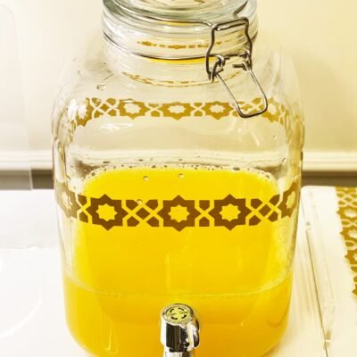 Glass beverage dispenser with yellow juice, sealed with a metal latch. It features a decorative geometric pattern and a chrome spigot, on a white table.