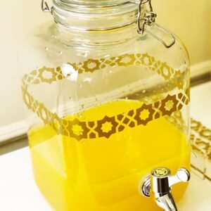 A clear glass drink dispenser with ornate patterns containing bright yellow juice and a shiny chrome spigot on a white surface, evoking freshness.