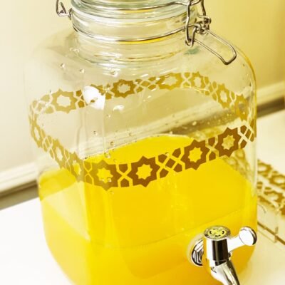 A clear glass drink dispenser with ornate patterns containing bright yellow juice and a shiny chrome spigot on a white surface, evoking freshness.