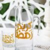Clear glass pitcher and tumbler with golden Arabic script on a white table. Background features green leaves and white geometric patterns.