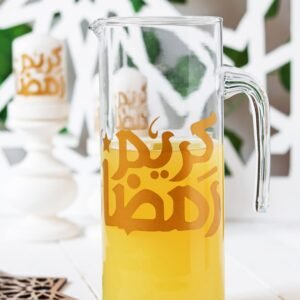 Glass pitcher with orange juice on white table, featuring Arabic script. Background includes geometric decor and a candle, evoking a festive mood.