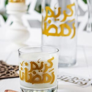 A glass of milk featuring gold Arabic script sits near dates on a white surface. In the background, a matching jug and candle create a serene Ramadan setting.