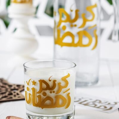 A glass of milk featuring gold Arabic script sits near dates on a white surface. In the background, a matching jug and candle create a serene Ramadan setting.