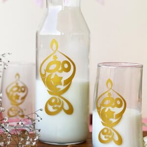 Glass jug and glass with milk, adorned with intricate gold Arabic calligraphy. Set on a wooden surface with dates and baby's breath flowers, creating a festive, elegant ambiance.