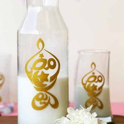 A glass pitcher and glass with milk, adorned with golden Arabic calligraphy, sit on a wooden table. A white flower and date are nearby, with a festive backdrop.