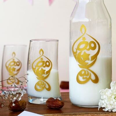 A milk-filled bottle and glasses with gold Arabic calligraphy on a wooden tray. Flowers and dates create a festive, elegant atmosphere.