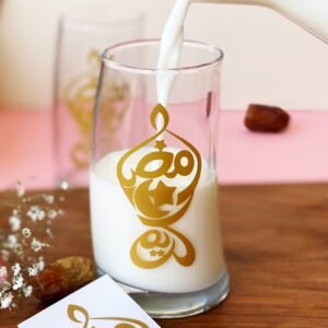 A glass of milk is being poured, featuring gold Arabic calligraphy. It sits on a wooden surface with dates and flowers, creating a warm, inviting scene.