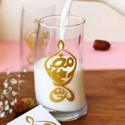 A glass of milk is being poured, featuring gold Arabic calligraphy. It sits on a wooden surface with dates and flowers, creating a warm, inviting scene.
