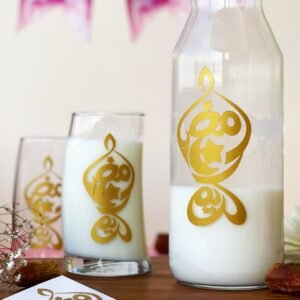 A carafe and two glasses filled with milk, adorned with gold Arabic calligraphy, placed on a wooden tray. In the background, festive banners hang, conveying a celebratory tone.
