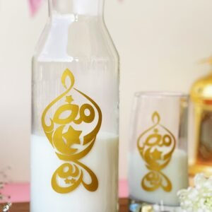 A glass pitcher and glass with white liquid, featuring gold Ramadan-themed Arabic calligraphy. Dates and white flowers are nearby on a wooden surface.