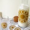 A glass carafe filled with milk and a matching glass, both featuring a gold geometric star pattern. Nearby are a cork lid and striped cloth.