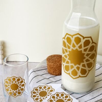 A glass carafe filled with milk and a matching glass, both featuring a gold geometric star pattern. Nearby are a cork lid and striped cloth.