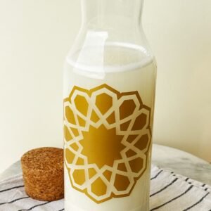 A clear glass carafe filled with milk, adorned with a golden geometric pattern, sits on a striped cloth next to a cork stopper, evoking a minimalist, elegant feel.