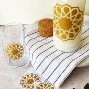 A glass bottle and tumbler with intricate gold star patterns sit on a striped cloth next to a cork stopper. Nearby, plant leaves add a fresh touch.