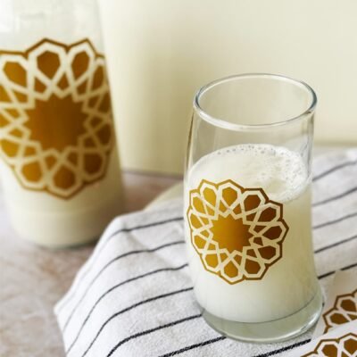 A glass and bottle with a creamy drink are adorned with gold geometric patterns, placed on a striped cloth. The setting feels calm and elegant.