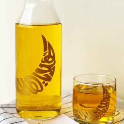 A tall glass carafe and a small glass tumbler filled with golden liquid, adorned with crescent moon designs, sit on a striped cloth beside green leaves.