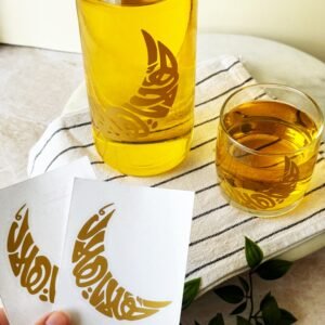 A bottle and glass of golden liquid with gold crescent moon designs sit on a striped cloth. Hand holds cards with matching designs. Green foliage nearby.