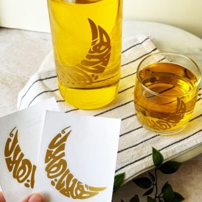 A bottle and glass of golden liquid with gold crescent moon designs sit on a striped cloth. Hand holds cards with matching designs. Green foliage nearby.