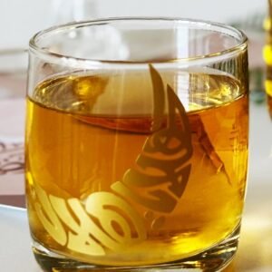 A glass of amber liquid with ornate golden Arabic calligraphy. The setting is bright and minimal, creating an elegant and serene atmosphere.