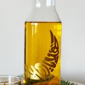 A clear glass carafe filled with amber liquid, featuring intricate gold Arabic calligraphy. A glass nearby contains the same drink. The scene is minimal and elegant.