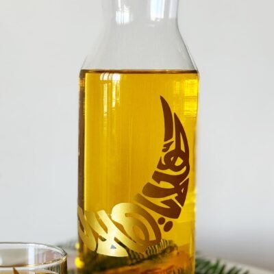 A clear glass carafe filled with amber liquid, featuring intricate gold Arabic calligraphy. A glass nearby contains the same drink. The scene is minimal and elegant.