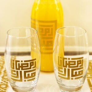 Two clear glasses and a bottle of orange juice, all adorned with gold geometric Arabic script, sit on a decorative table, conveying a festive atmosphere.