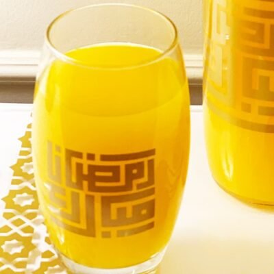 A glass of vibrant orange juice with geometric gold patterns sits on a table beside a similarly decorated bottle. Bright and inviting atmosphere.