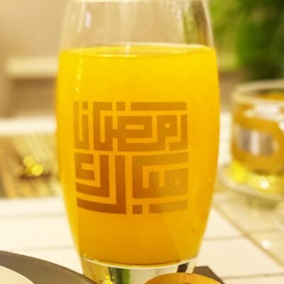 A glass of orange juice with Arabic calligraphy on the side is placed on a table. Next to it is a gold spoon and a black plate. The setting is warm and inviting.