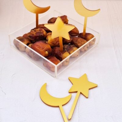 A clear box filled with dates is decorated with gold crescent and star sticks. Two sticks are placed on a white surface, creating a festive feel.