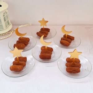 Six glass plates with syrupy pastries topped with gold crescent moon and star picks. A white lantern and pearl beads sit nearby on a pastel surface.