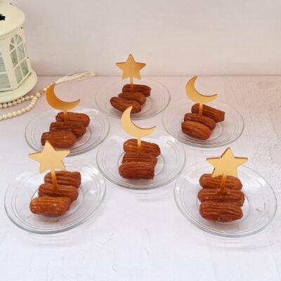 Six glass plates with syrupy pastries topped with gold crescent moon and star picks. A white lantern and pearl beads sit nearby on a pastel surface.