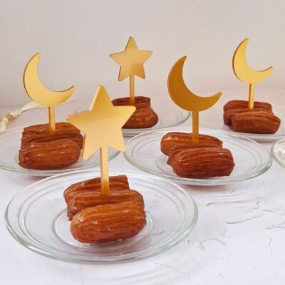 Small glass plates with syrupy pastries are adorned with golden star and crescent moon toppers. The display is festive and elegantly arranged on a white surface.