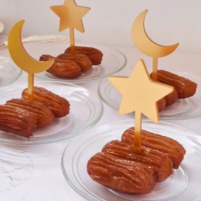 Three glass plates, each with two syrupy pastries, are topped with golden star and crescent moon decorations, creating a festive, elegant display.