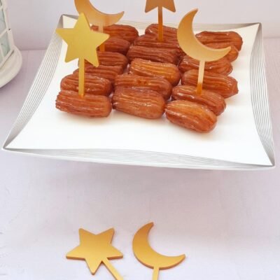 Golden-brown pastries on a white plate, decorated with star and crescent-shaped toppers. The setting is elegant and festive.