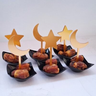 Black dishes with dates topped by gold crescent moons and stars on toothpicks, arranged neatly on a white surface; festive and elegant presentation.