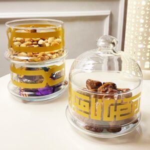 Elegant glass jars with golden designs hold nuts, chocolates, and dates on a table. The setting is warm and inviting, reflecting a festive atmosphere.