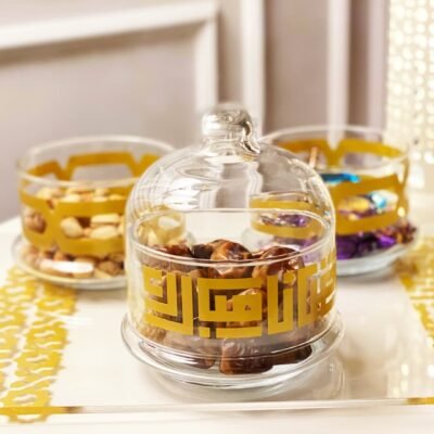 Glass jars on a white table hold various snacks like dates and nuts. The jars have golden Arabic script, evoking a warm, festive atmosphere.