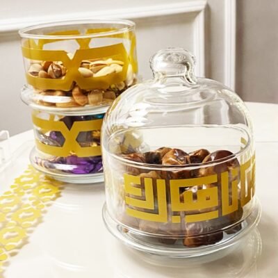 Glass jars with gold geometric patterns sit on a white tray, containing assorted nuts and dates. The setting is elegant and inviting.